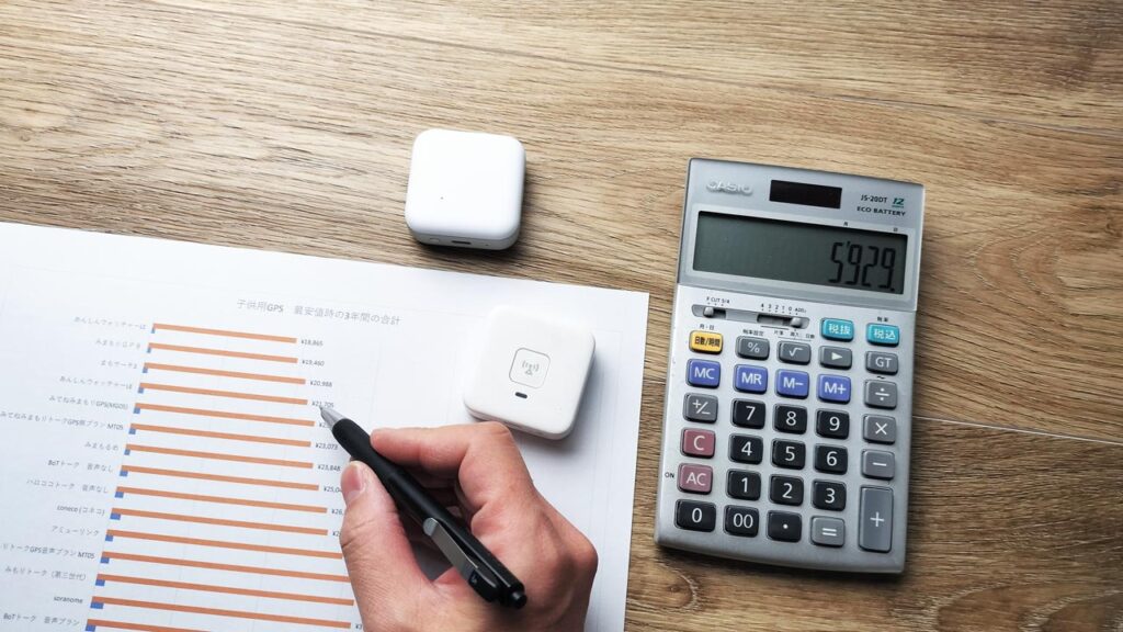 ノートにあんしんウォッチャーLEとみまもりGPSそれぞれの本体代と月額料金を書き出し、家計への負担を比較している保護者の写真。