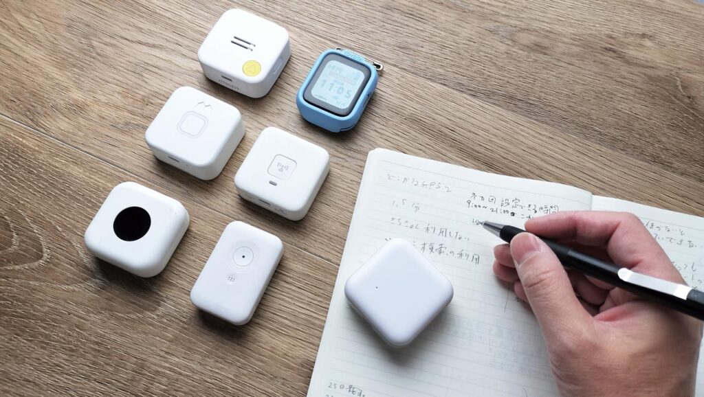 机の上に子ども用GPS端末がいくつか並び、ノートとペン、スマートフォンが置かれている写真。こどもGPS研究所・所長もふが精度を検証中
