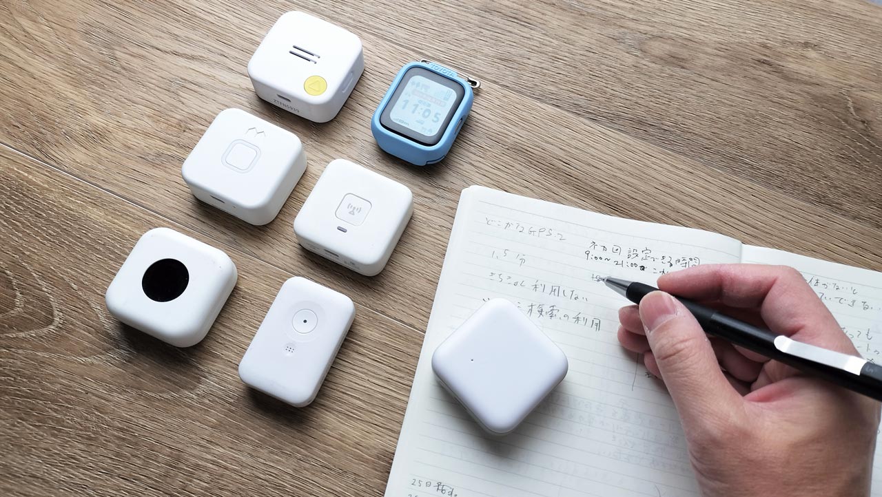 机の上に子ども用GPS端末がいくつか並び、ノートとペン、スマートフォンが置かれている写真。こどもGPS研究所・所長もふが精度を検証中