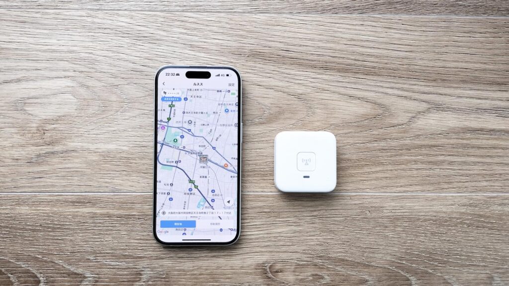 あんしんウォッチャーLEの子どもGPS端末と、バッテリー残量と位置情報が表示されたスマホ画面を並べた写真。