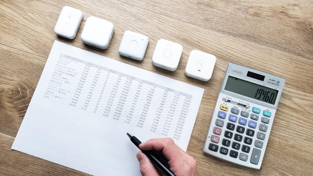 5機種の子どもGPSを並べ、その下に料金比較表が置かれているイメージ図。3年トータルコストを見比べている様子。