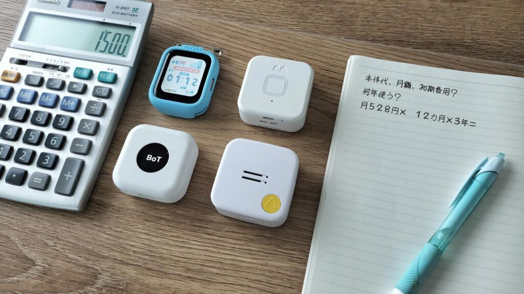 ノートに「本体代」「月額」「初期費用」など料金の項目が手書きされ、その横にこども用GPS端末が置かれている写真。
