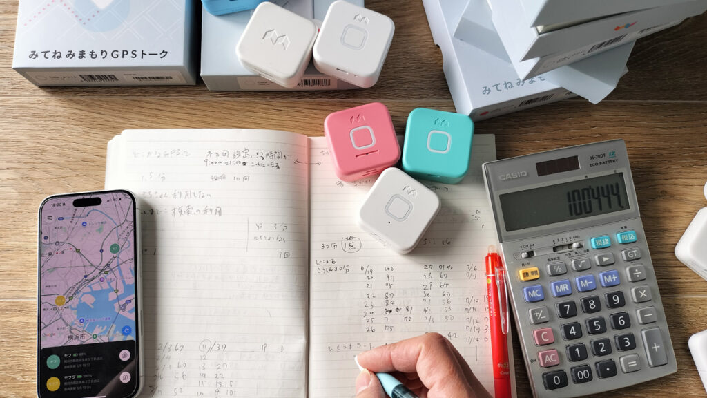机の上に子ども用GPS端末がいくつか並び、ノートとペン、スマートフォンが置かれている写真。こどもGPS研究所・所長もふが子どもGPSを検証しているイメージ。